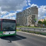 Jadący ulicą autobus miejski linii 34. Obok i dalej w tle widoczne bloki mieszkalne, znaki drogowe i drzewa. Niebo zachmurzone.