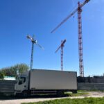 Biały samochód ciężarowy stojący obok placu budowy. Na owym placu stoją trzy żurawie wieżowe. W tle drzewa.